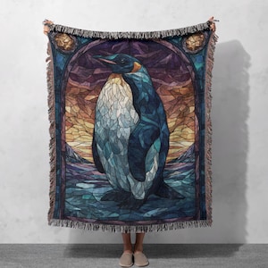 May include: A woven throw blanket featuring a central penguin illustration rendered in a stained-glass style. The design incorporates shades of blue, purple, and orange, framed by an arched border with fringed edges on the top and bottom sides.