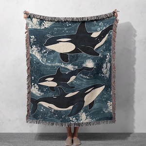 May include: A woven throw blanket with a blue background and a pattern of three black and white orcas swimming in the ocean. The orcas are surrounded by bubbles and seaweed.
