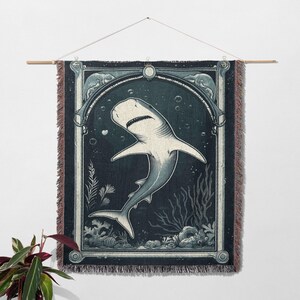 May include: A woven wall tapestry featuring a white shark swimming in a dark blue ocean scene. The shark is depicted in a vintage style with intricate details. The tapestry has a dark blue background with a light blue border.