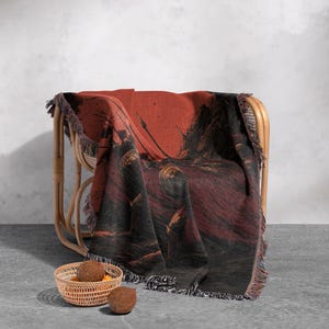 May include: A woven throw blanket draped over a wicker chair. The blanket features a dark red and black illustration with fringe trim. A small woven basket with fruit sits in front of the chair.