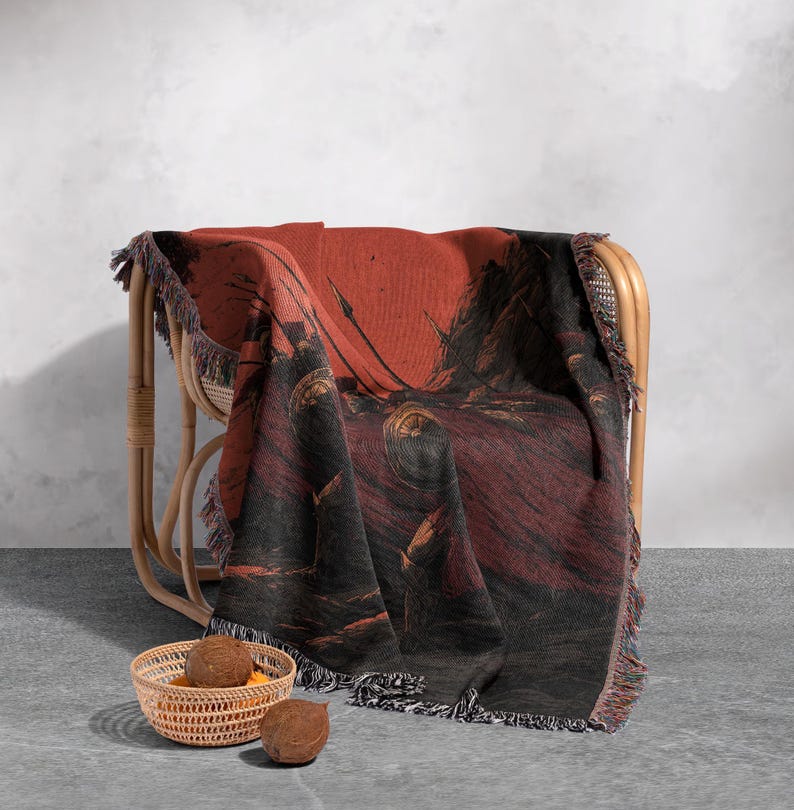 May include: A woven throw blanket draped over a wicker chair. The blanket features a dark red and black illustration with fringe trim. A small woven basket with fruit sits in front of the chair.