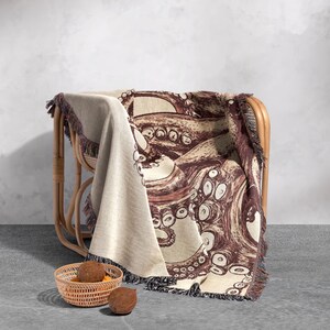 May include: A woven throw blanket with a brown octopus design on a cream background. The blanket is draped over a wicker chair and has fringe on the edges.
