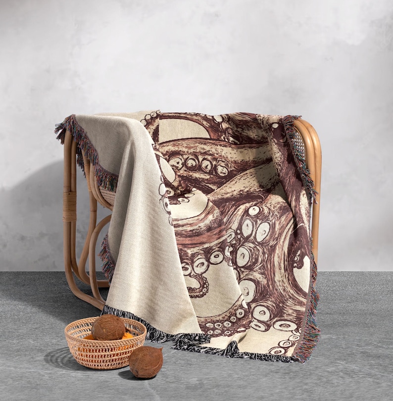 May include: A woven throw blanket with a brown octopus design on a cream background. The blanket is draped over a wicker chair and has fringe on the edges.