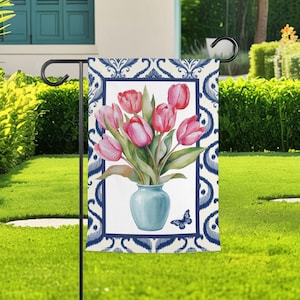 Peut inclure: Un drapeau de jardin rectangulaire présentant une illustration centrale de tulipes roses dans un vase bleu clair avec un petit papillon bleu à côté. Le motif est encadré par un damas bleu foncé et blanc sur fond blanc, suspendu à un support.
