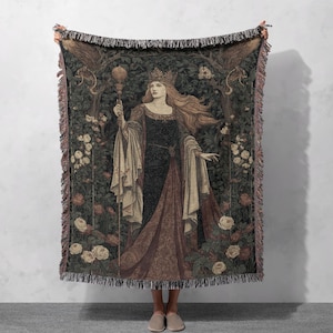 May include: A tapestry throw blanket featuring a medieval queen holding a scepter, wearing a crown, and standing in a garden. The throw has a dark green background with roses and trees, and a fringed edge.