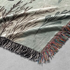 May include: A woven throw blanket with a green background and a black and white floral pattern. The blanket has a fringe border with a multi-colored yarn.
