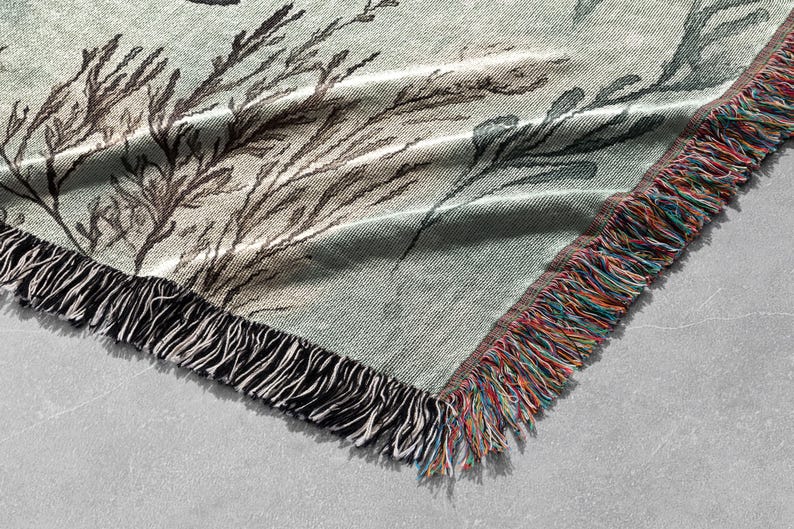 May include: A woven throw blanket with a green background and a black and white floral pattern. The blanket has a fringe border with a multi-colored yarn.