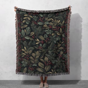 May include: A dark green throw blanket with a botanical pattern of leaves and red berries. The blanket has a fringed edge and is held up against a neutral background. The design features various shades of green and brown.