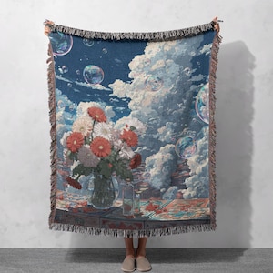 May include: A woven throw blanket featuring a dreamy landscape with a vase of flowers, bubbles, and a cloudy sky. The color palette includes shades of blue, white, red, and brown. The blanket has a fringed edge.
