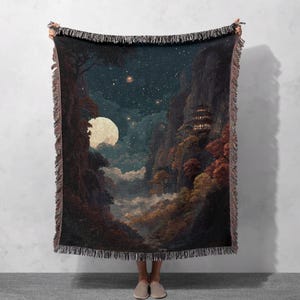 May include: A woven throw blanket featuring a nighttime landscape with a full moon, stars, and a pagoda nestled in a mountain valley. The color palette includes deep blues, browns, and oranges. The blanket has a fringed edge.