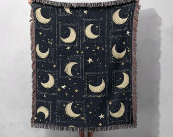 May include: A dark blue throw blanket featuring a repeating pattern of gold crescent moons and stars within square frames. The blanket has a fringed edge and is held up against a neutral background.
