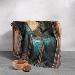 May include: A woven throw blanket draped over a wicker chair. The blanket features a teal and black forest scene with bats and a full moon. The blanket has a fringed edge and is paired with a small woven basket and two coconuts.