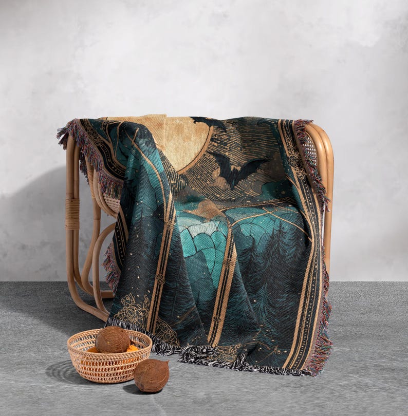 May include: A woven throw blanket draped over a wicker chair. The blanket features a teal and black forest scene with bats and a full moon. The blanket has a fringed edge and is paired with a small woven basket and two coconuts.