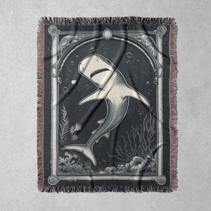 May include: A black and white woven tapestry featuring a stylized illustration of a shark swimming in the ocean. The shark is depicted in a vintage style with intricate details. The tapestry has a fringed border.
