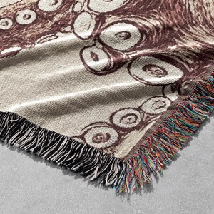 May include: A brown and white woven throw blanket featuring an octopus design. The blanket has a fringe border with multicoloured threads.