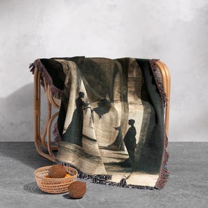 May include: A woven throw blanket with a black and white silhouette design of two figures in a hallway. The blanket has a fringe border and is draped over a wicker chair.