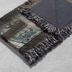 Pu&ograve; includere: Una coperta tessuta piegata con un disegno di vetrata. La coperta ha un retro grigio scuro ed &egrave; rifinita con frange bianche e nere. L'immagine sulla coperta include sfumature di blu, verde e marrone.