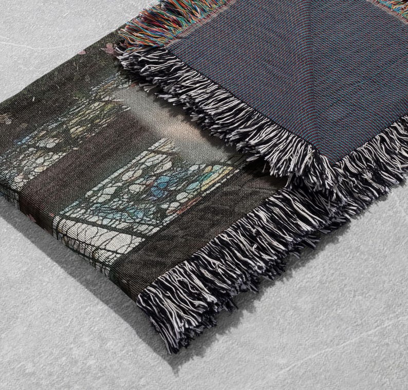 Pu&ograve; includere: Una coperta tessuta piegata con un disegno di vetrata. La coperta ha un retro grigio scuro ed &egrave; rifinita con frange bianche e nere. L'immagine sulla coperta include sfumature di blu, verde e marrone.