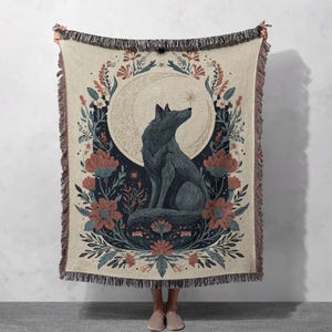Peut inclure: Un plaid tissé présentant un loup bleu foncé central assis de profil devant un grand croissant de lune crème. Le motif est encadré par des illustrations botaniques complexes de fleurs rouges et bleues et de feuillage sur un fond beige clair.