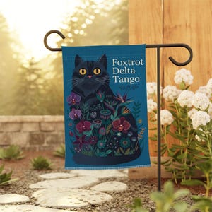 May include: A rectangular garden flag with a blue background featuring a black cat surrounded by colorful flowers and the text Foxtrot Delta Tango. The flag is suspended from a black metal stand, displaying a detailed illustration of a cat.