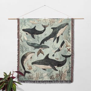May include: A woven wall tapestry featuring a blue-green background with multiple black whales swimming in a stylized ocean scene. The tapestry has a fringed border.
