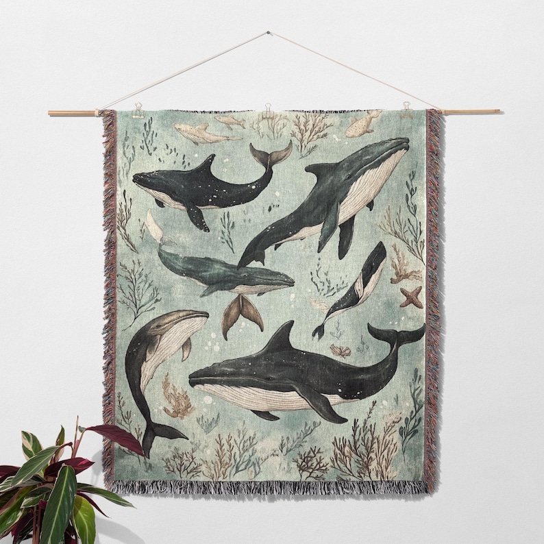 May include: A woven wall tapestry featuring a blue-green background with multiple black whales swimming in a stylized ocean scene. The tapestry has a fringed border.