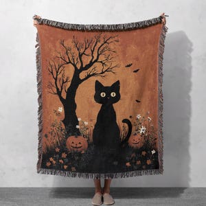 Könnte beinhalten: Eine gewebte Wurkdecke mit einem Halloween-Design. Sie zeigt eine schwarze Katze, Kürbisse und einen kahlen Baum auf orangefarbenem Hintergrund. Das Design umfasst Fledermäuse und Blumen, und die Decke hat einen Fransenrand.