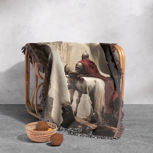 May include: A woven tapestry throw blanket features a medieval knight riding a white horse through a stone archway. The knight is wearing red armor and a red cape. The tapestry is brown, white, and red.