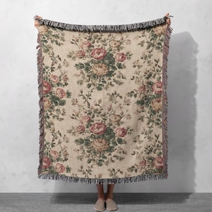 Op de afbeelding: Een beige bloemen plaid met een herhalend patroon van roze en groene rozen en bladeren. De plaid heeft een gefranjerde rand in zwart en bordeaux. De plaid wordt tegen een witte muur gehouden.