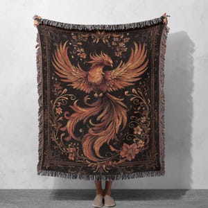 May include: A woven tapestry throw blanket featuring a detailed illustration of a phoenix with orange and red feathers against a dark background with intricate floral designs.