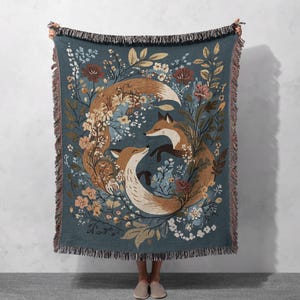 May include: A teal throw blanket featuring a circular design of two foxes surrounded by flowers and foliage. The foxes are brown and white, with the flowers in shades of cream, peach, and brown. The blanket has a fringed edge.