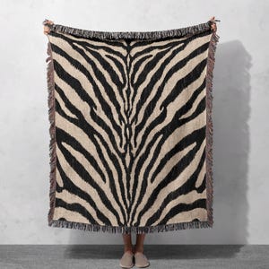 May include: A rectangular woven throw blanket featuring a high-contrast black and beige zebra stripe pattern. The textile has a textured appearance with visible woven threads and decorative fringed edges along all four sides of the fabric.