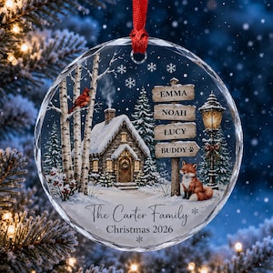 Peut inclure: Une décoration ronde en acrylique transparent illustrant une scène hivernale avec un chalet enneigé, des bouleaux, un cardinal rouge, un renard et un lampadaire. Un panneau en bois indique les noms Emma, Noah, Lucy et Buddy. Texte : The Carter Family Christmas 2026.