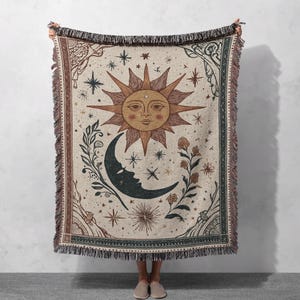 May include: A beige woven throw blanket featuring a sun, moon, and star design. The sun has a face and is surrounded by rays. The moon has a face and is dark blue. The blanket has a fringed edge and a decorative border.