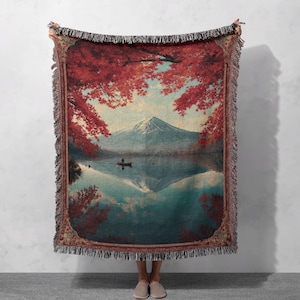 May include: Woven throw blanket featuring a scenic landscape with a mountain, lake, and red foliage. The design includes a boat on the water and a mountain reflected in the lake. The blanket has a fringed edge.