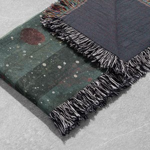 May include: A folded blanket with a dark teal and gray color scheme. The blanket features a pattern of a large red circle and white dots on the teal side. The edges are finished with black and white fringe.