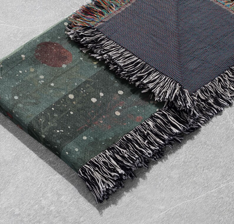 May include: A folded blanket with a dark teal and gray color scheme. The blanket features a pattern of a large red circle and white dots on the teal side. The edges are finished with black and white fringe.