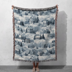 Puede incluir: Una manta tejida azul y blanca con una escena de un pueblo invernal con casas, árboles y colinas nevadas. La manta tiene un borde con flecos en tonos marrones y azules. El diseño es un patrón repetido.