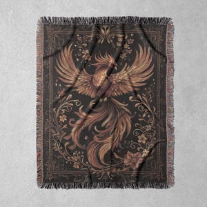 May include: A woven tapestry throw blanket featuring a gold phoenix with intricate details against a dark background. The phoenix is surrounded by floral designs and a decorative border.