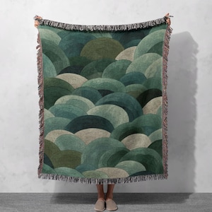 May include: A woven throw blanket featuring a repeating pattern of overlapping, semi-circular shapes in shades of green and beige. The blanket has a fringed edge and is held up against a white wall.