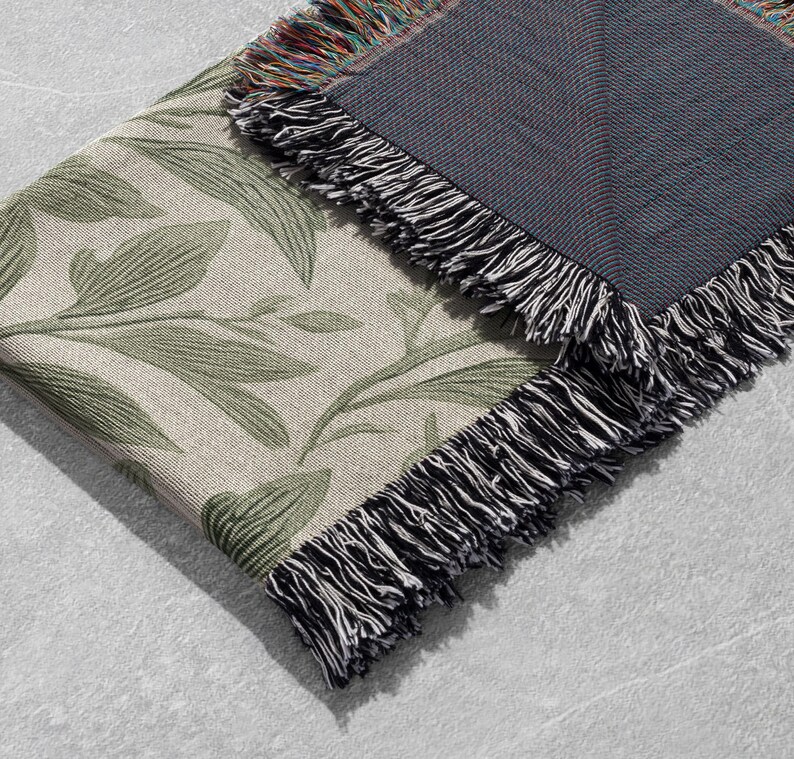 Op de afbeelding: Een geweven sprei met een botanisch bladerpatroon in groen en beige. De sprei heeft een donkergrijze achterkant en een franjeborduur in zwart en wit. De sprei is opgevouwen en toont het patroon en de franjes.