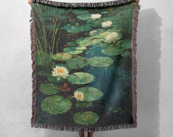 May include: A woven throw blanket depicting a tranquil pond scene with water lilies. The design features green lily pads, white flowers, and a dark blue water background. The blanket has a fringed edge and is held up by a person.