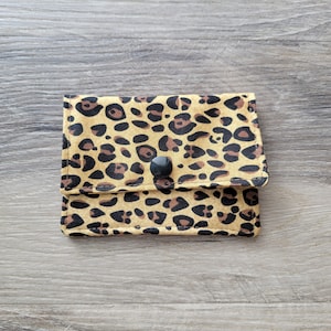 May include: A small, square wallet with a leopard print pattern in shades of tan, brown, and black. The wallet has a flap closure secured by a black button. The fabric appears to be cotton or a similar material.