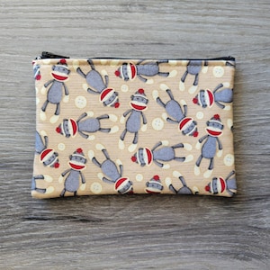 May include: A beige zippered pouch with a repeating pattern of gray sock monkeys wearing red hats. The monkeys are in various poses and have white accents on their feet.