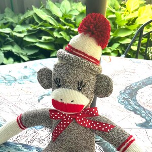 May include: A handmade sock monkey doll with a red bow tie, hat, and pom-pom. The monkey is gray with white accents on the arms and legs, and red stripes. The doll is sitting on a patterned table.