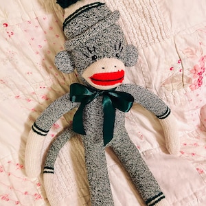 May include: A handmade sock monkey doll with a gray and white knitted body, white limbs, and a red mouth. It wears a matching hat with a dark green pom-pom and a green bow. The doll is lying on a floral patterned quilt.