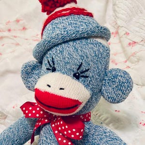 May include: A handmade sock monkey doll with a blue and white knit body, a red and white polka dot bow, and a red pom-pom hat. The monkey has a white face, red lips, and black eyelashes.