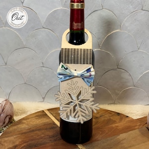 May include: A wooden wine bottle holder with a snowflake design. A bow tie made of money is attached to the holder. The bottle holder is wrapped around a bottle of red wine.