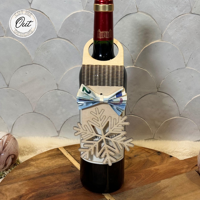 May include: A wooden wine bottle holder with a snowflake design. A bow tie made of money is attached to the holder. The bottle holder is wrapped around a bottle of red wine.