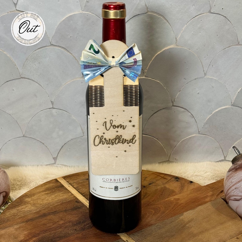May include: A wooden wine bottle tag with a bow made from folded bills. The tag has the text "Vom Christkind" and gold glitter stars.